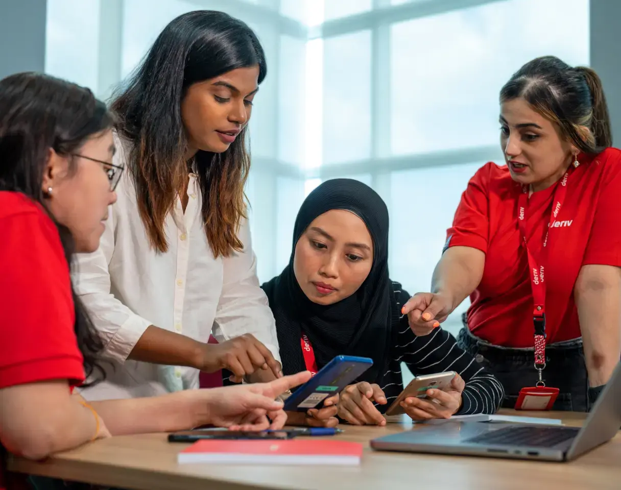 A collaborative team discussion at Deriv, with members sharing ideas and reviewing information on mobile devices and a laptop.